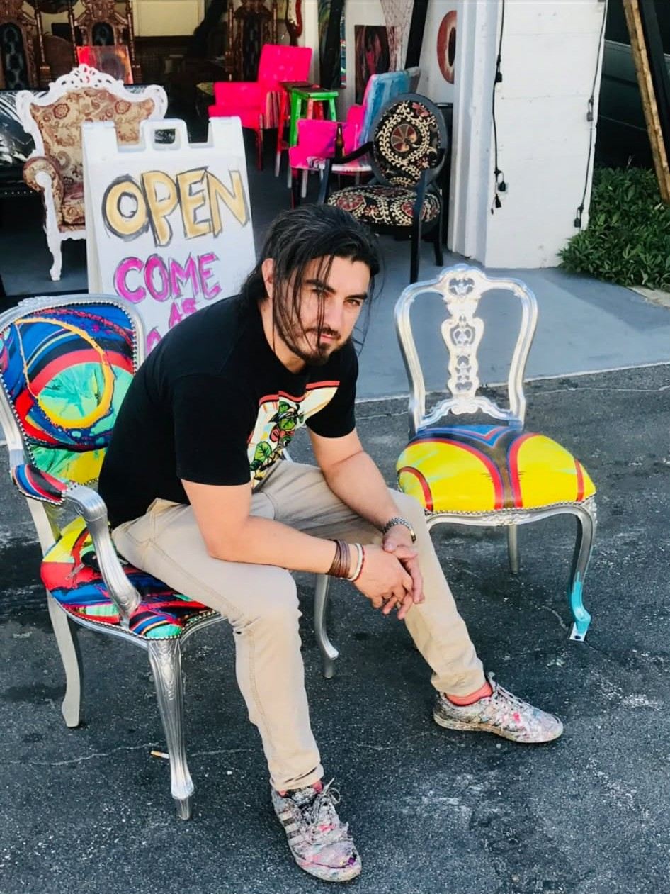 Nicolas Bogado seated between two hand-painted chairs outside his Lake Worth studio